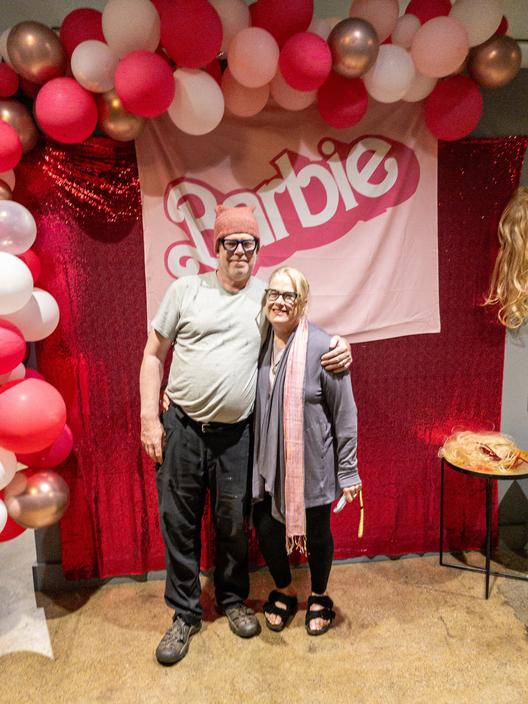 Man and woman standing in front of Barbie sign and balloons.