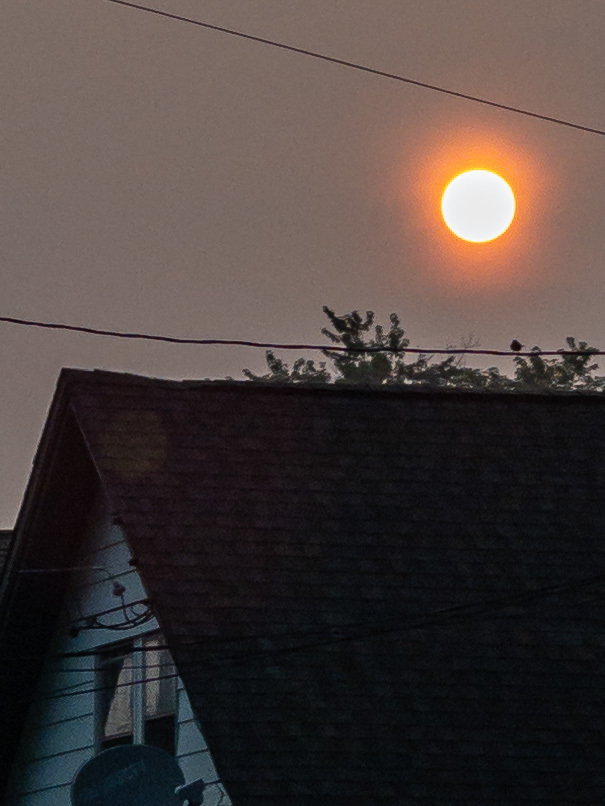 Closeup of sun rising above a house roof ridge line, dimmed by smoke haze from Canadian wildfires.