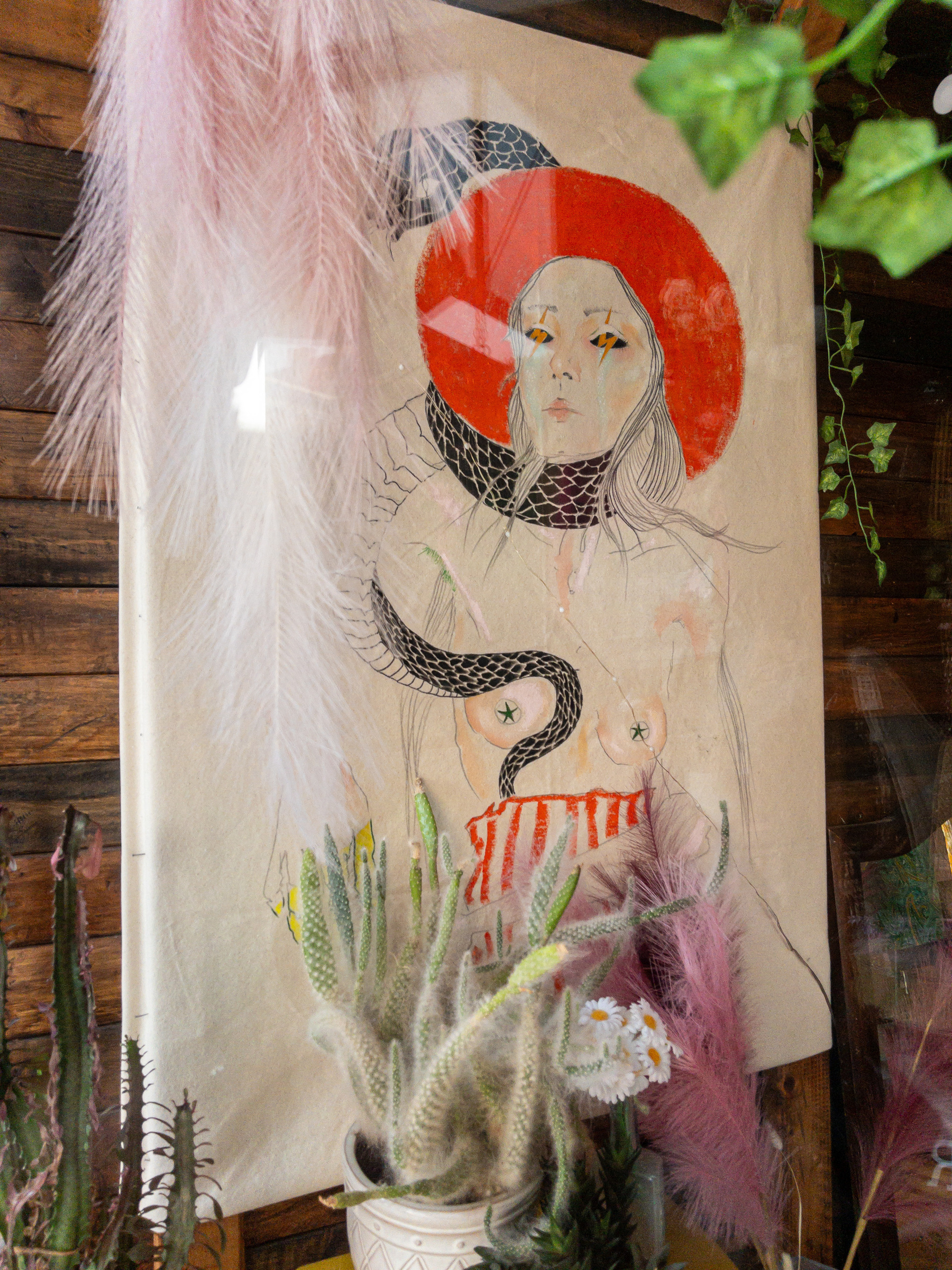 Stylized nude portrait of woman with bright, broad brimmed orange hat, snake wrapped around her neck, with tail wrapped around one breast, in a women’s second hand clothing shop window.