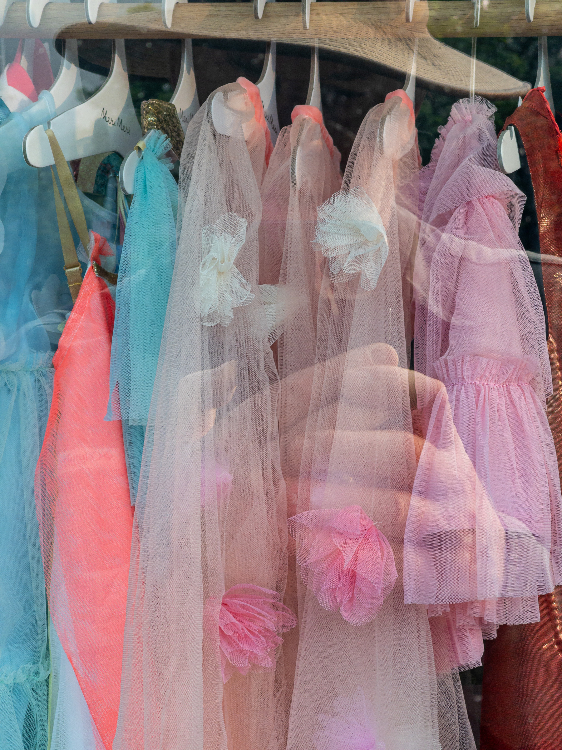 Pastel colored, gauzy children’s dresses hanging in a shop window with the reflection of the photographer in the window layered over the top.