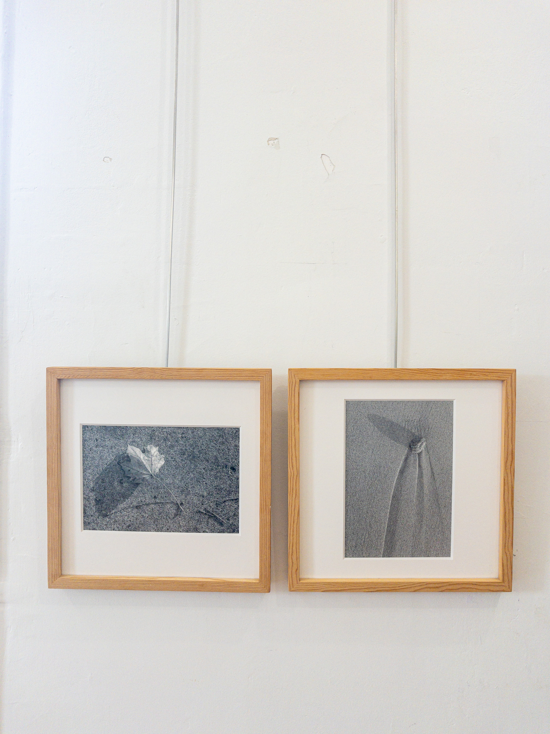 Two framed photographs at an exhibition depicting a leaf and shadow and a stone on the beach with erosion trails extending down from it.