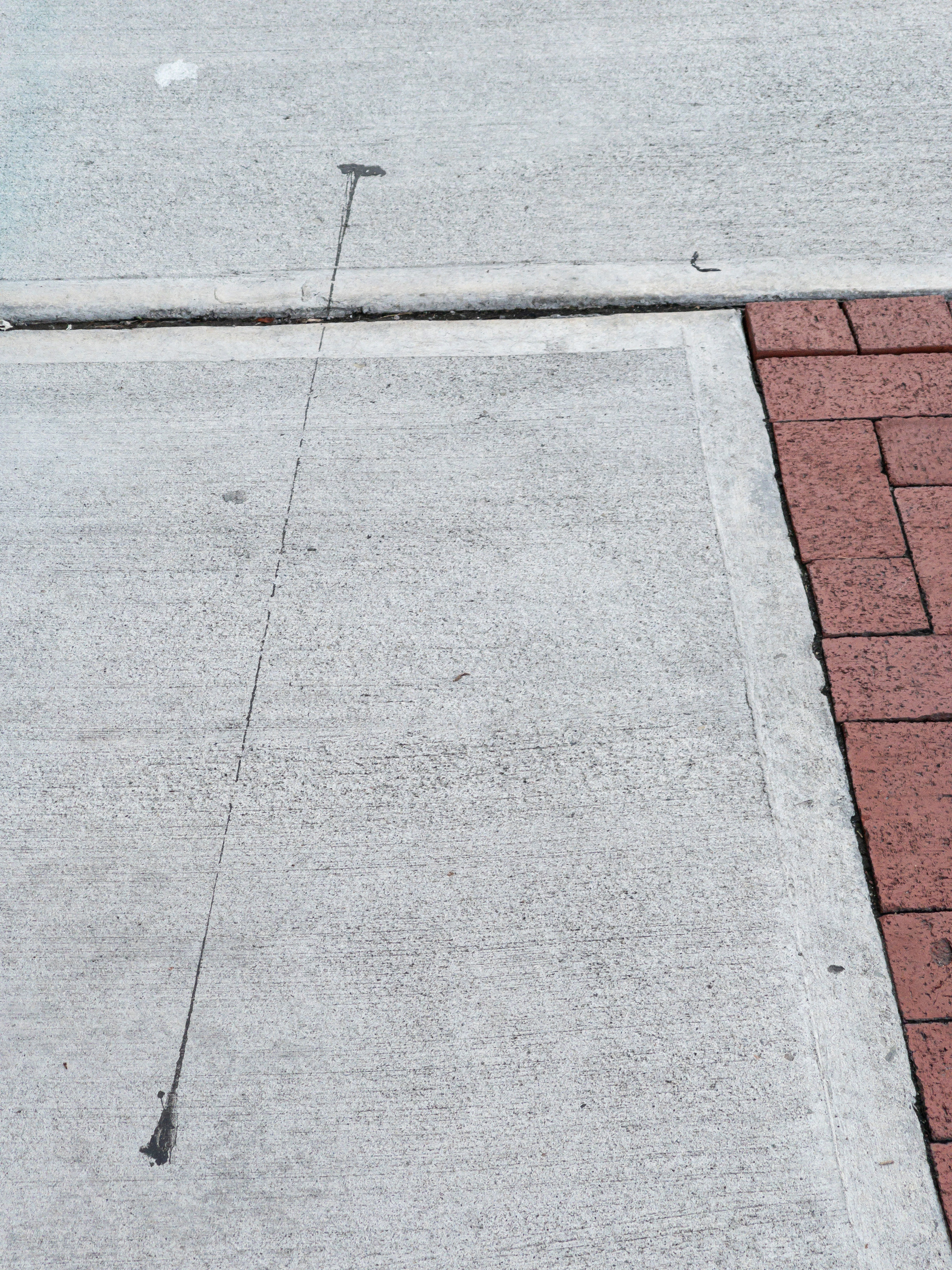 A black line of something tar like dribbled from the top middle of the frame to the bottom across an expansion joint in the concrete sidewalk. A triangle of brick paving to the right.