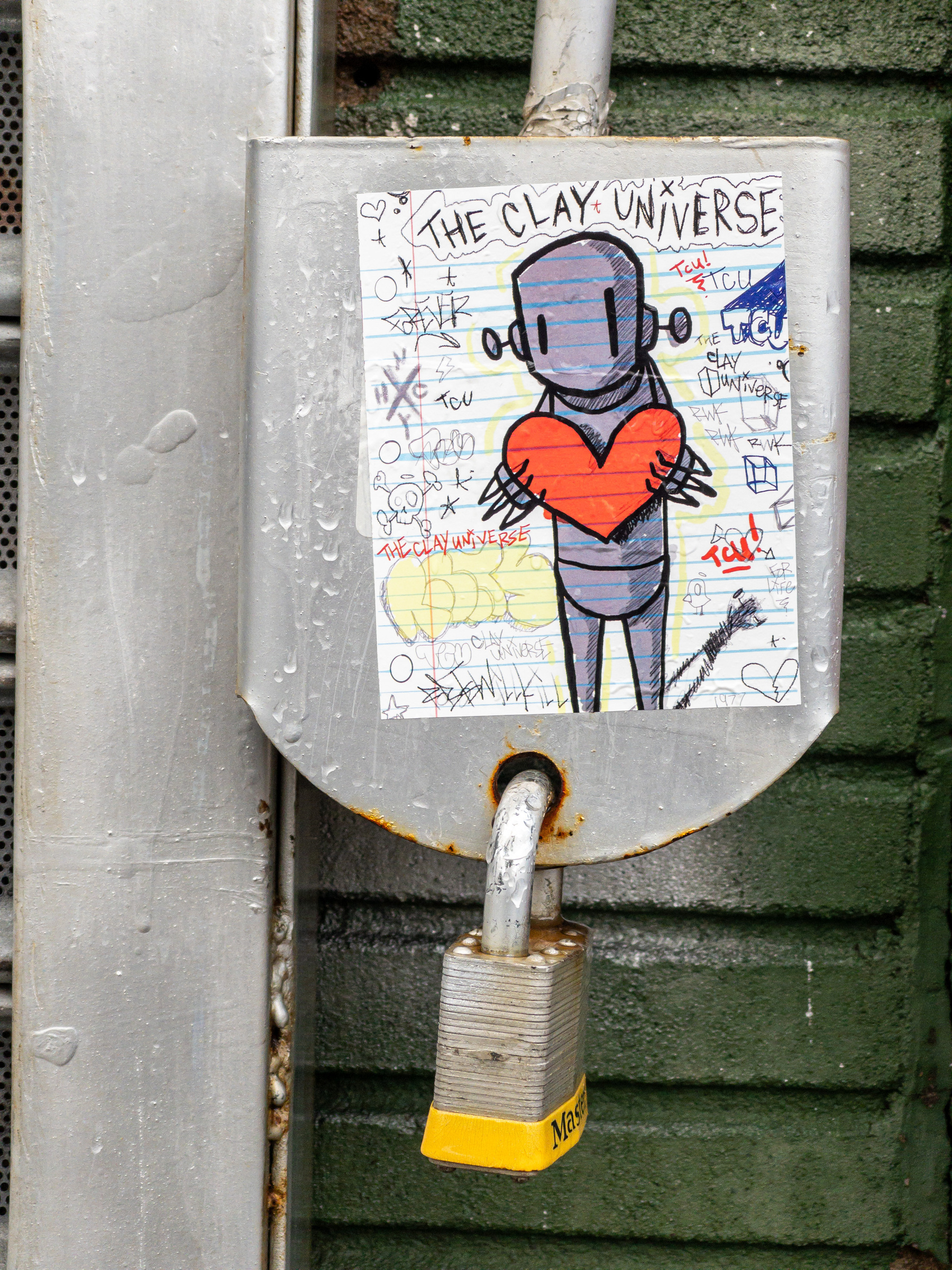 Sticker on a gate lock plate with yellow plastic bottomed lock. The sticker depicts a cartoon robot character holding a heart with “The Clay Universe” written above.