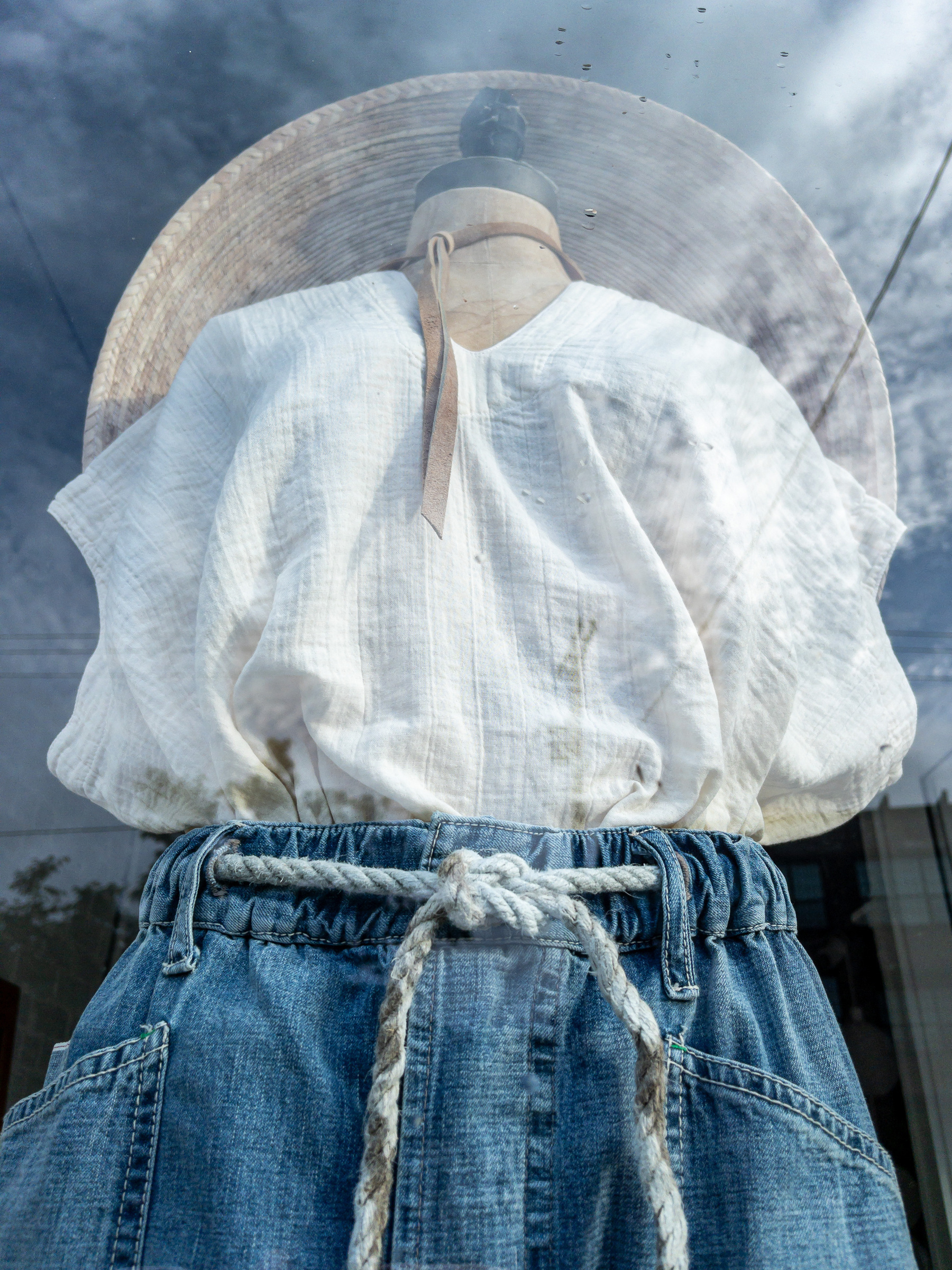 Blue jeans, rope belt, white linen blouse and broad brim hat in shop window with partially cloudy sky reflection overlaid.