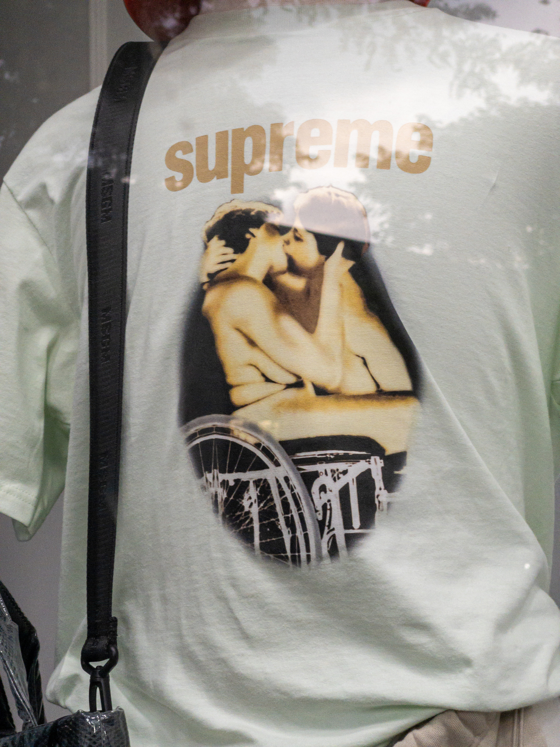 Closeup of the back of a white t-shirt with “supreme” written and a screen shot of a person of unknown sex kissing a woman sitting in a wheelchair. Both are naked.