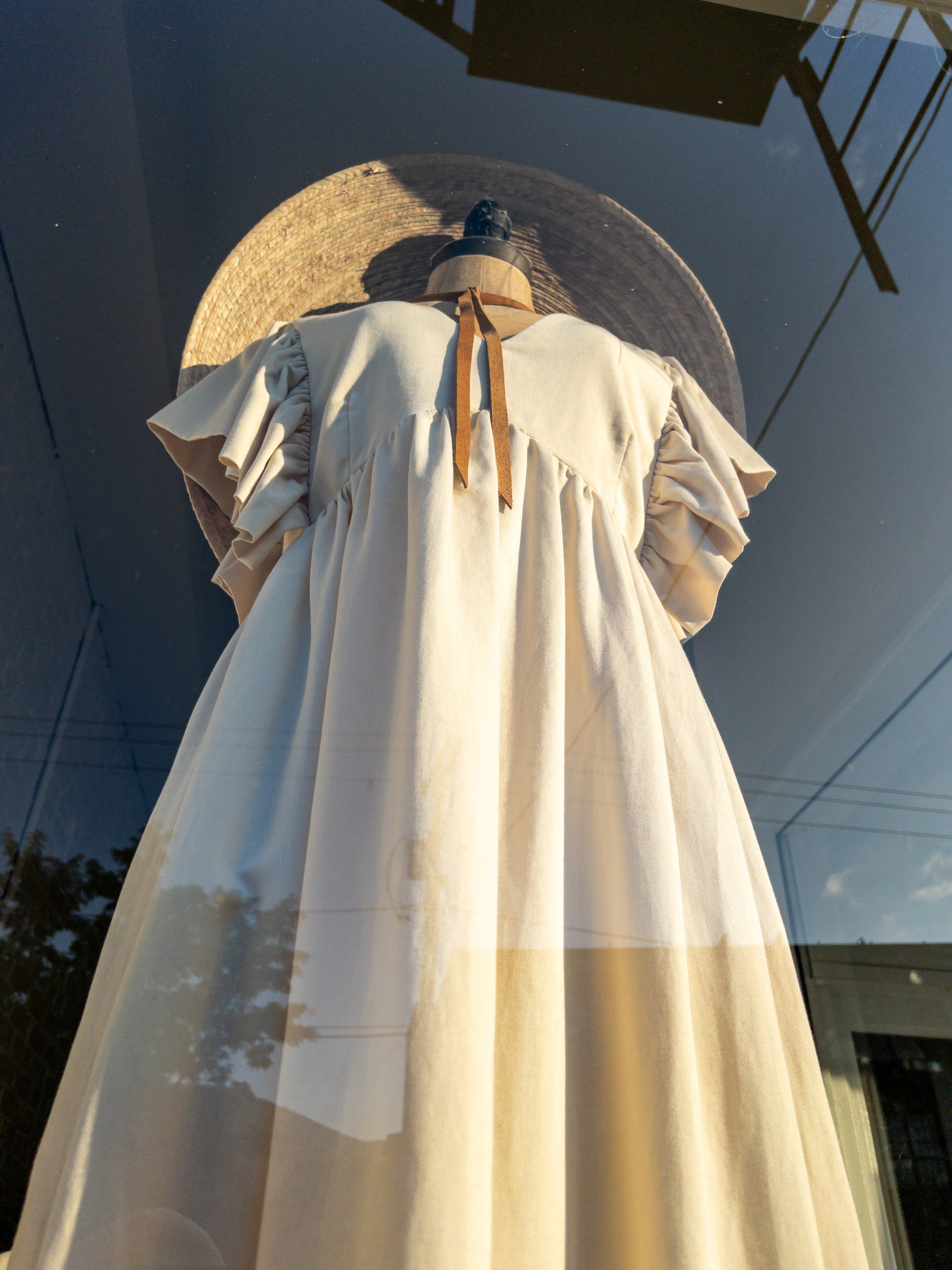 Off-white cotton dress on mannequin gathered under the bust line with pleated short sleeves. Large brim straw hat caps it off. Sky and buildings reflection overlaid.
