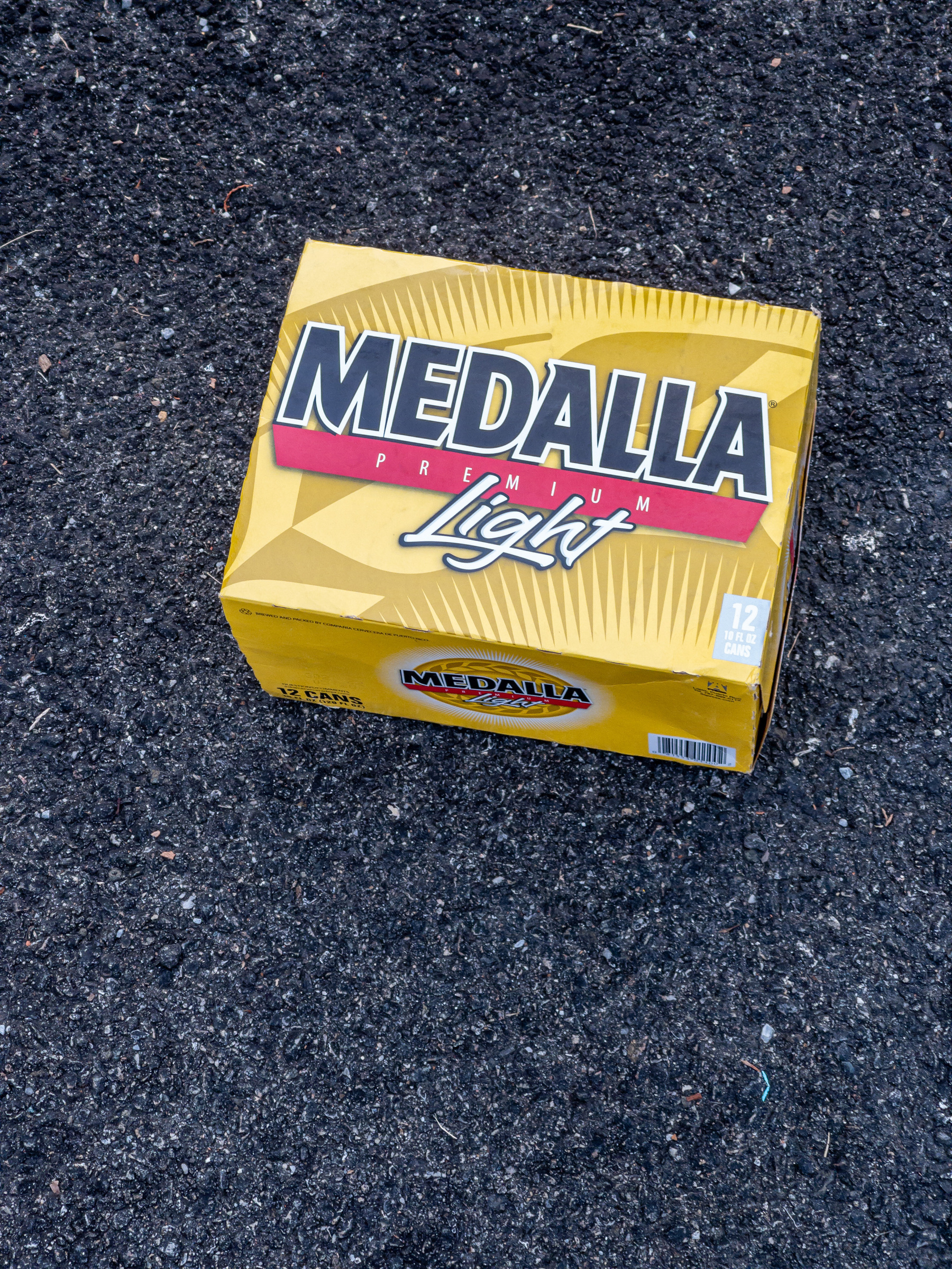 Medalla Premium Light 12 Can box on asphalt pavement. Box is shades of yellow leaf motif background with “light” in script letters, white with black outline. “Premium” is white sans serif lettering on red stripe. “Medalla” is navy blue stylized sans serif lettering with white outline above the red stripe.