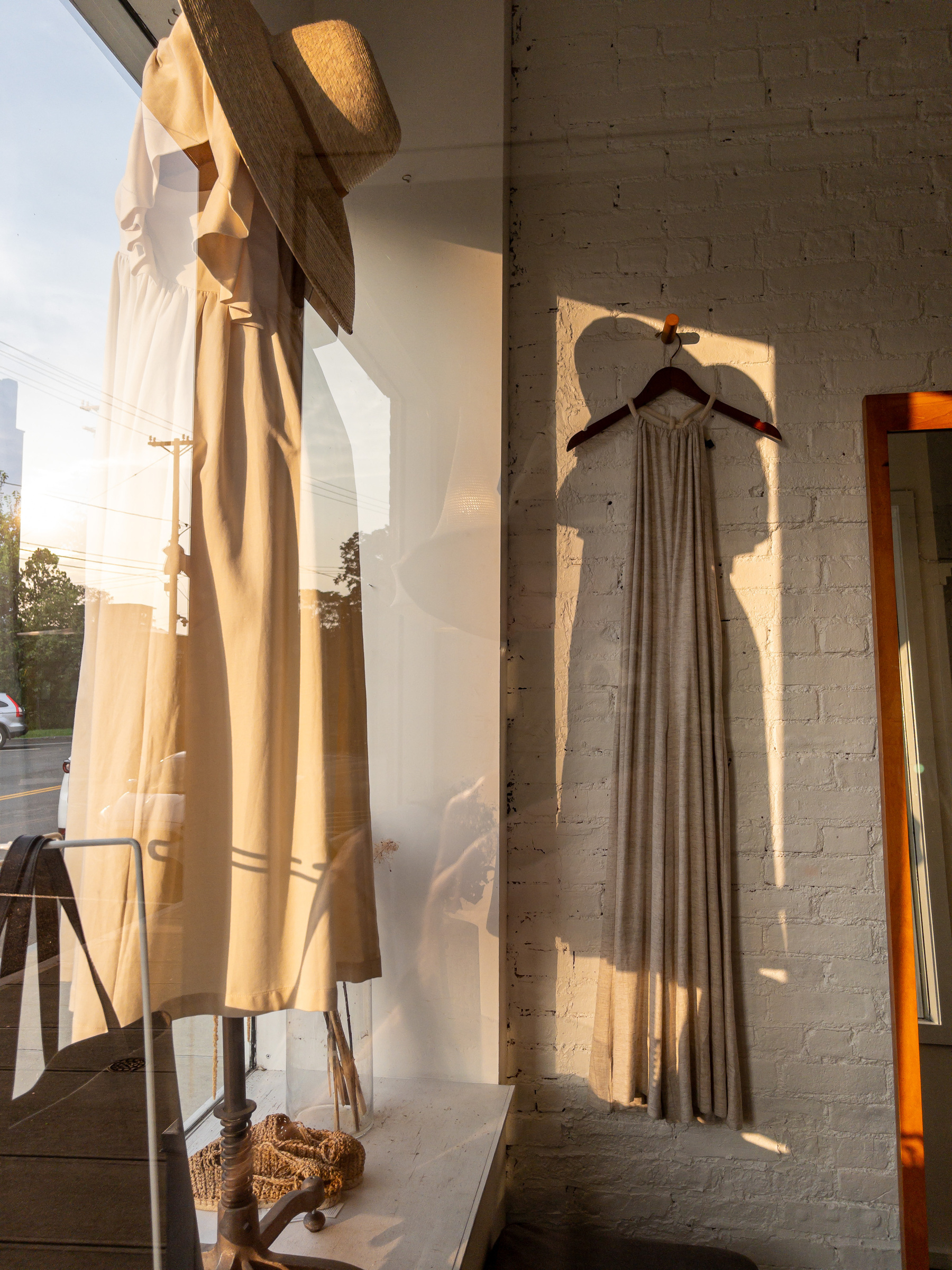 Interior of women’s clothing store with off white dress on a mannequin in the window and on the left and a sundress of similar color hanging on the wall from a hanger.