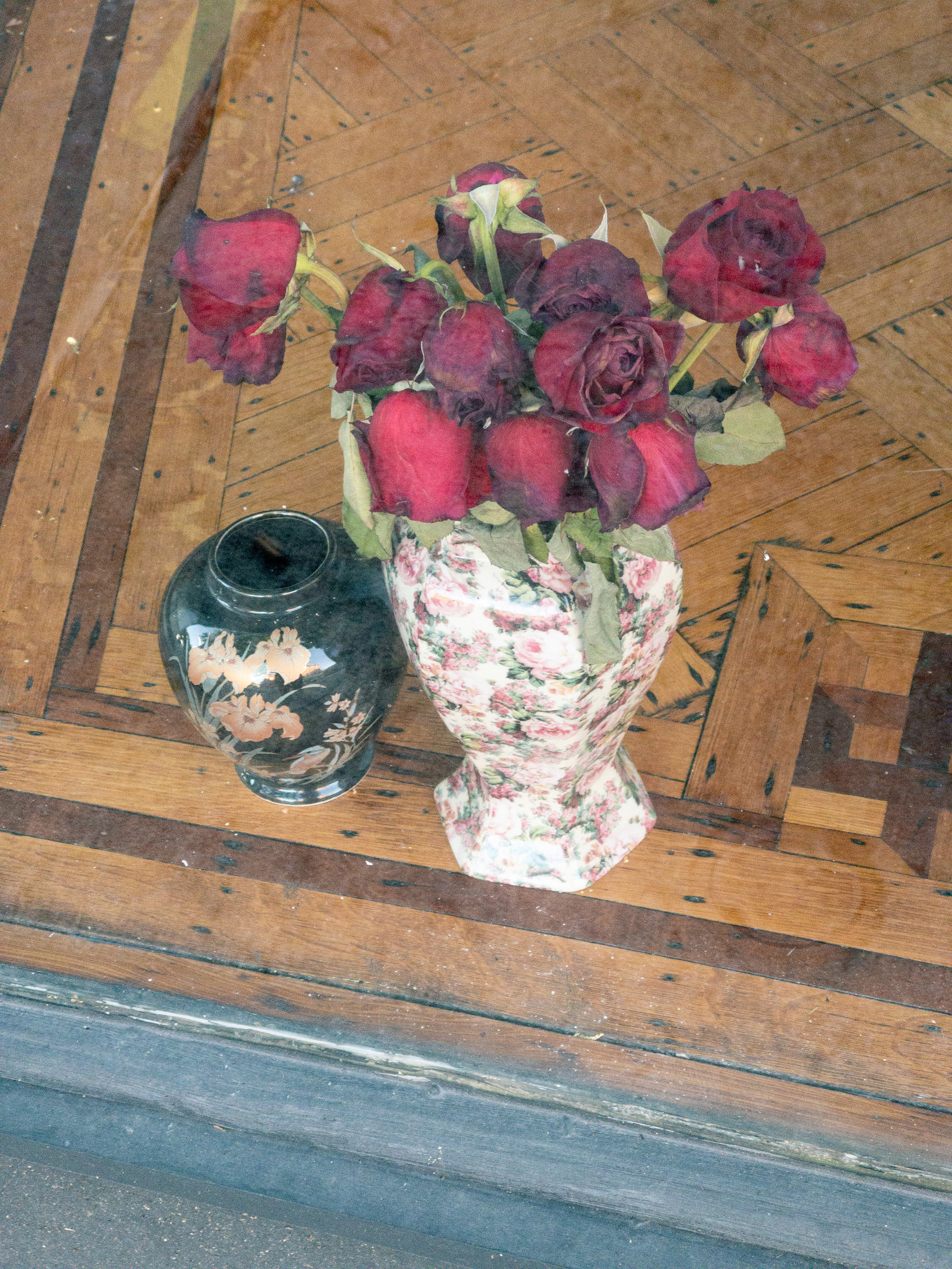 small vase and large floral pattern vase with wilting roses sitting on wood inlay display surface in a shop window