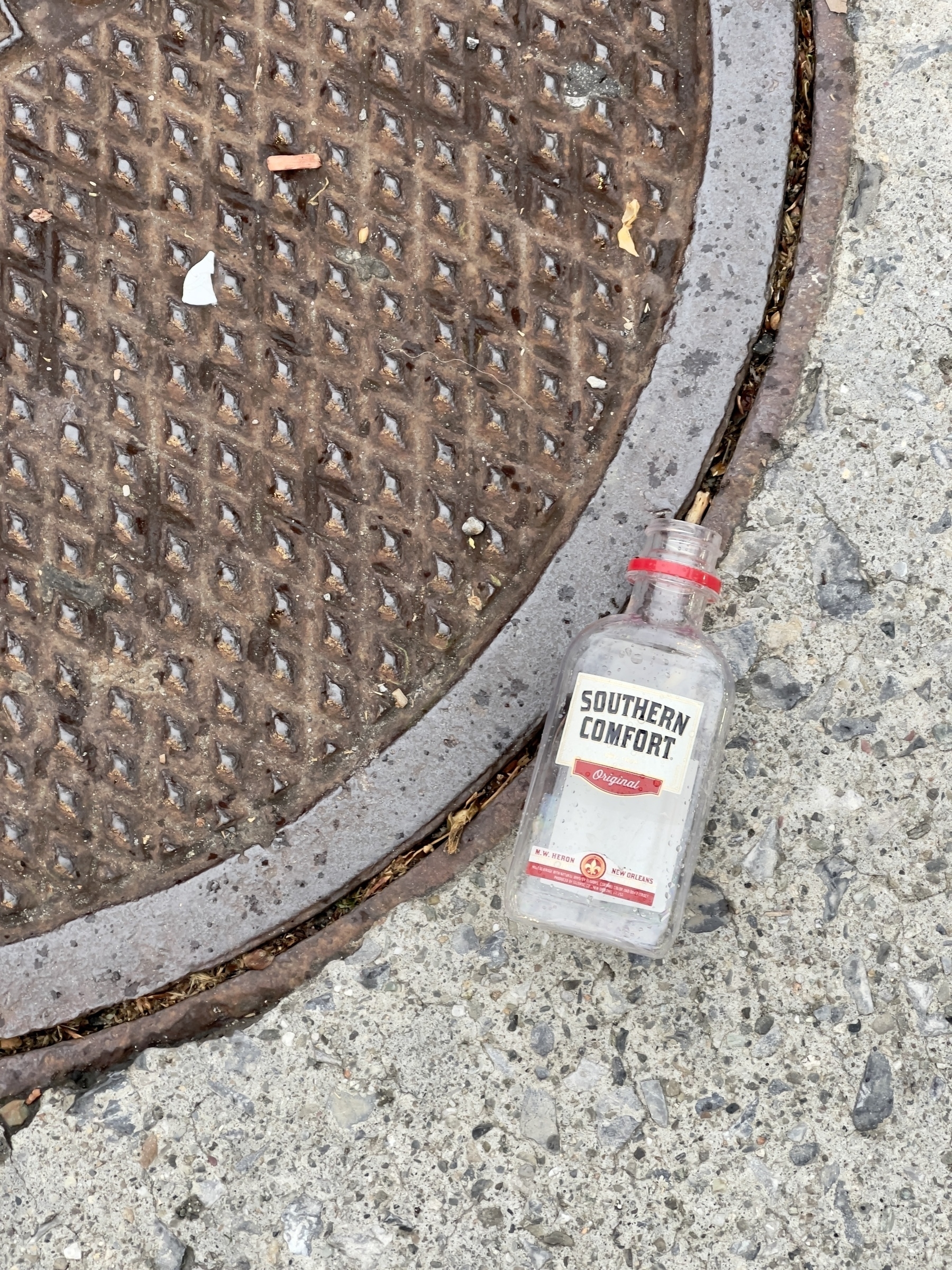 Mini Southern Comfort bottle near the edge of a manhole cover.