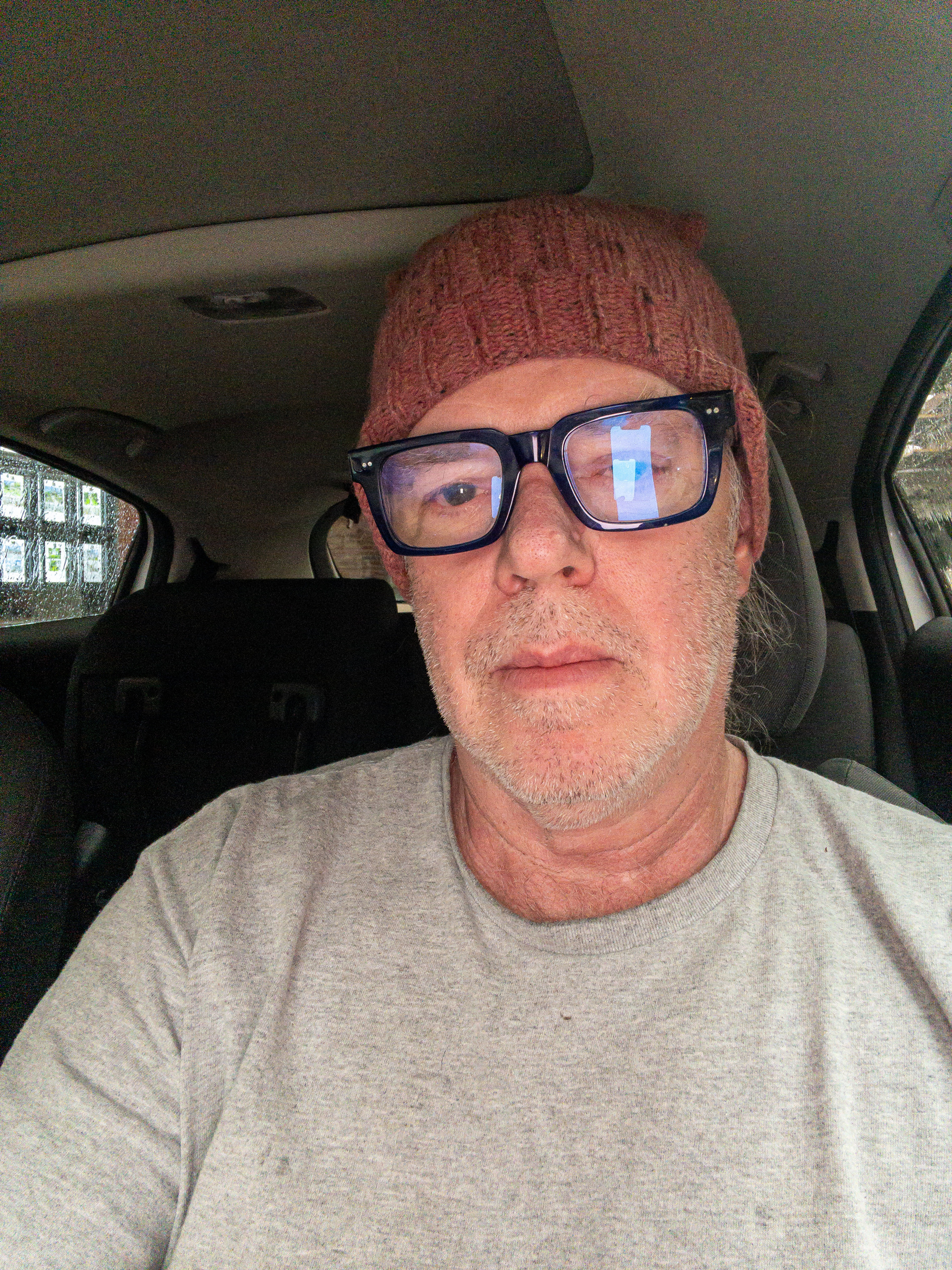 Selfie, man in a car with gray t-shirt, beard stubble, heavy framed blue glasses, pink knit cap.