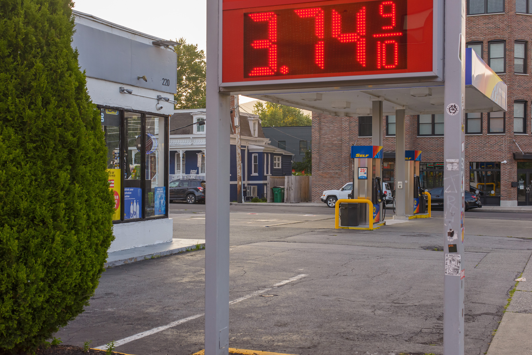 street scape, gas station, sign with gas prices in the middle foreground, evergreen shrub in the foreground on left side of frame, gas pumps in the background between the supports of the pricing sign, edge of convenience store in the background between the evergreen and left support of the pricing sign