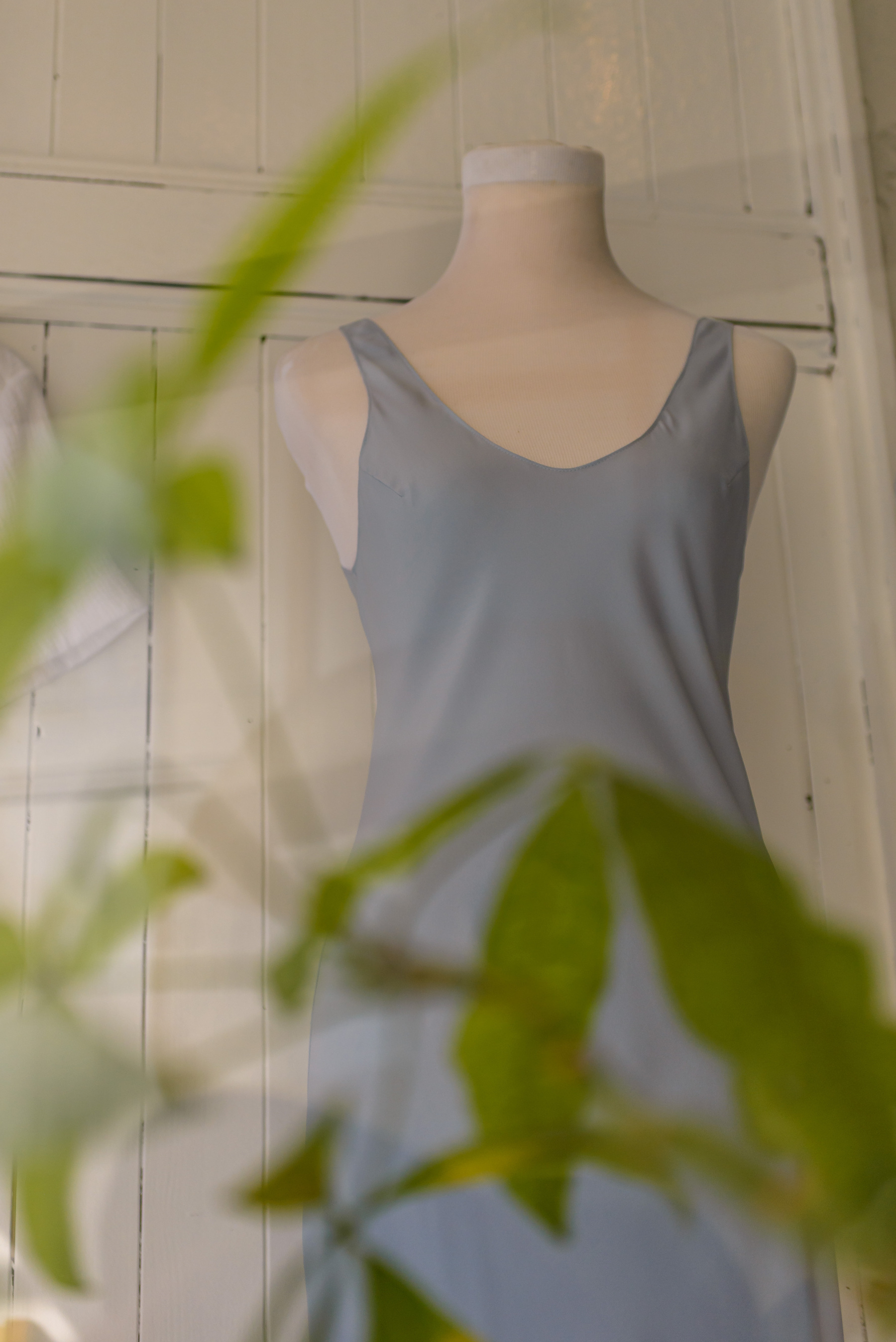 blue/gray silk sundress on a mannequin form with blurred leaves from a plant in front screening the lower part of the dress